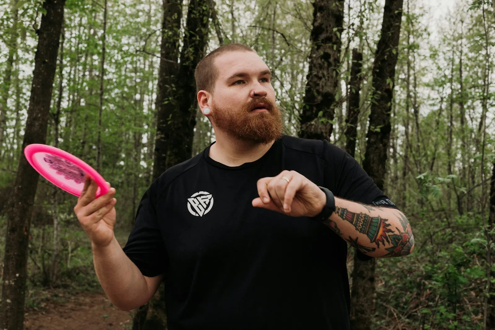 Disc golfer in a wooded course checking his wrist gear while holding a bright pink disc