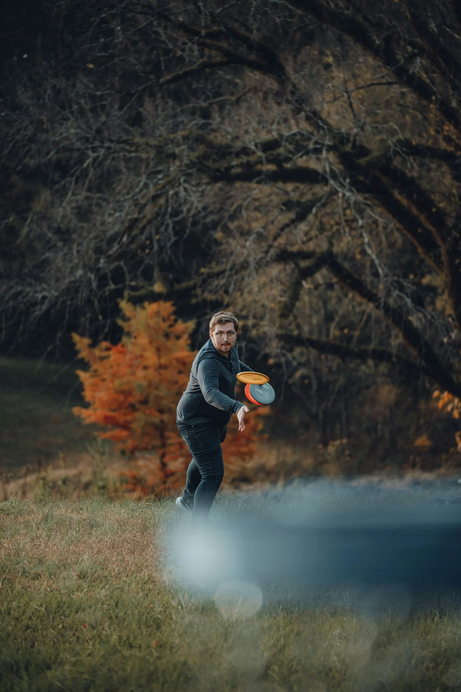 Disc golfer mid-throw launching a driver across an autumn fairway lined with trees