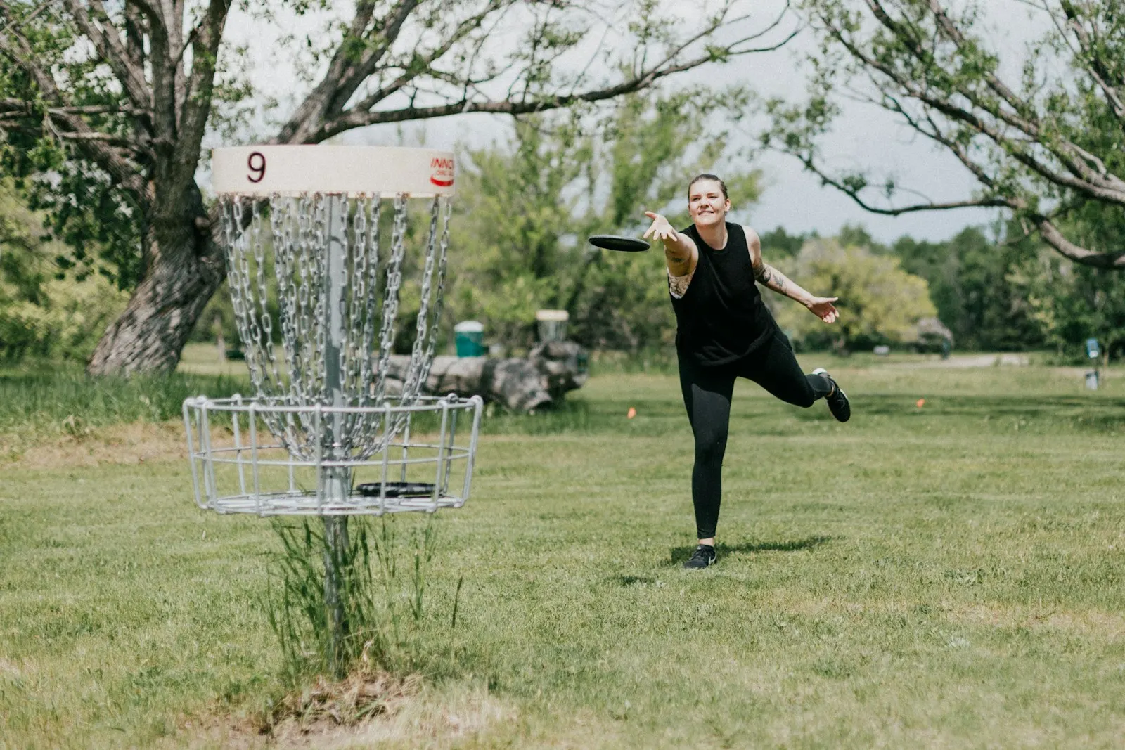 Disc golfer releasing a black disc toward a chain basket on an open grassy course