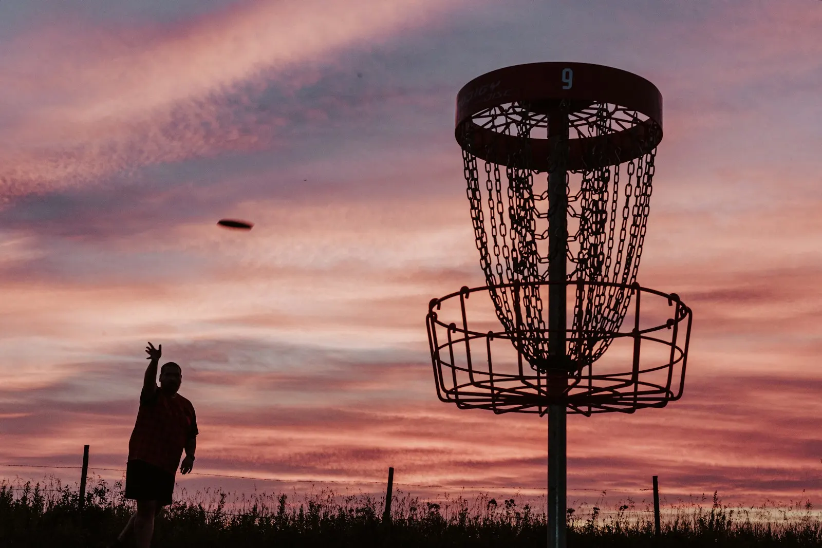 Silhouetted player following through on a long throw beside a basket against a pink sunset sky