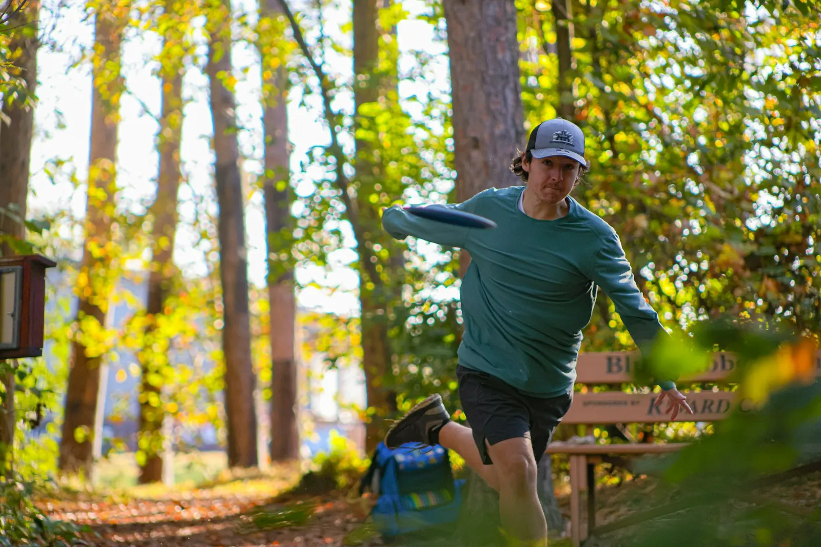 Player powering through a backhand throw on a sunlit wooded fairway with a blue disc in hand