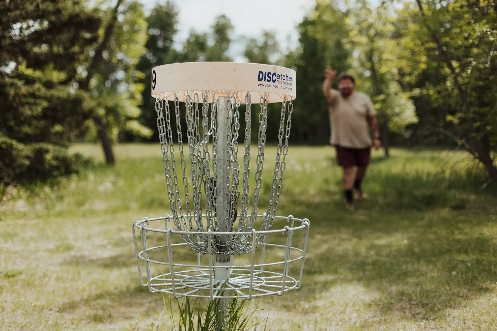 Close-up of a DISCatcher basket with a player approaching after a putt on a tree-lined course